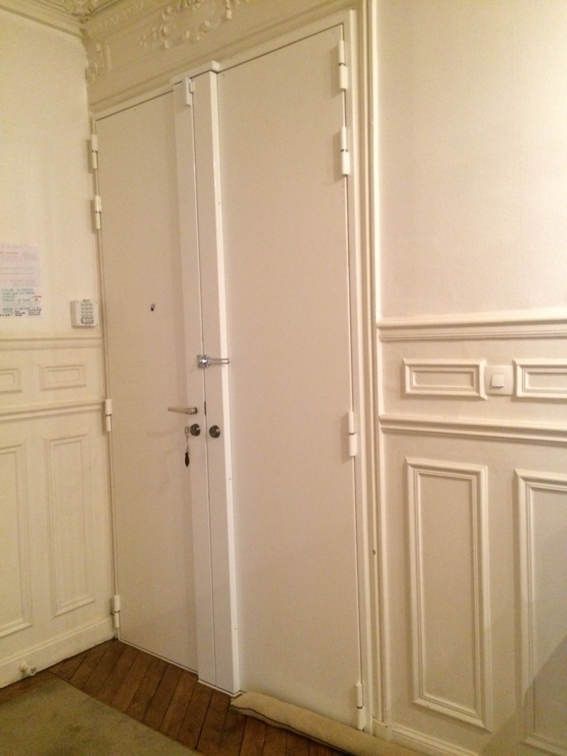 Porte d'entrée blanche à double vantail renforcée par un blindage, paumelles et cornières apparentes, dans un couloir d'immeuble ancien mouluré — Charenton-le-Pont