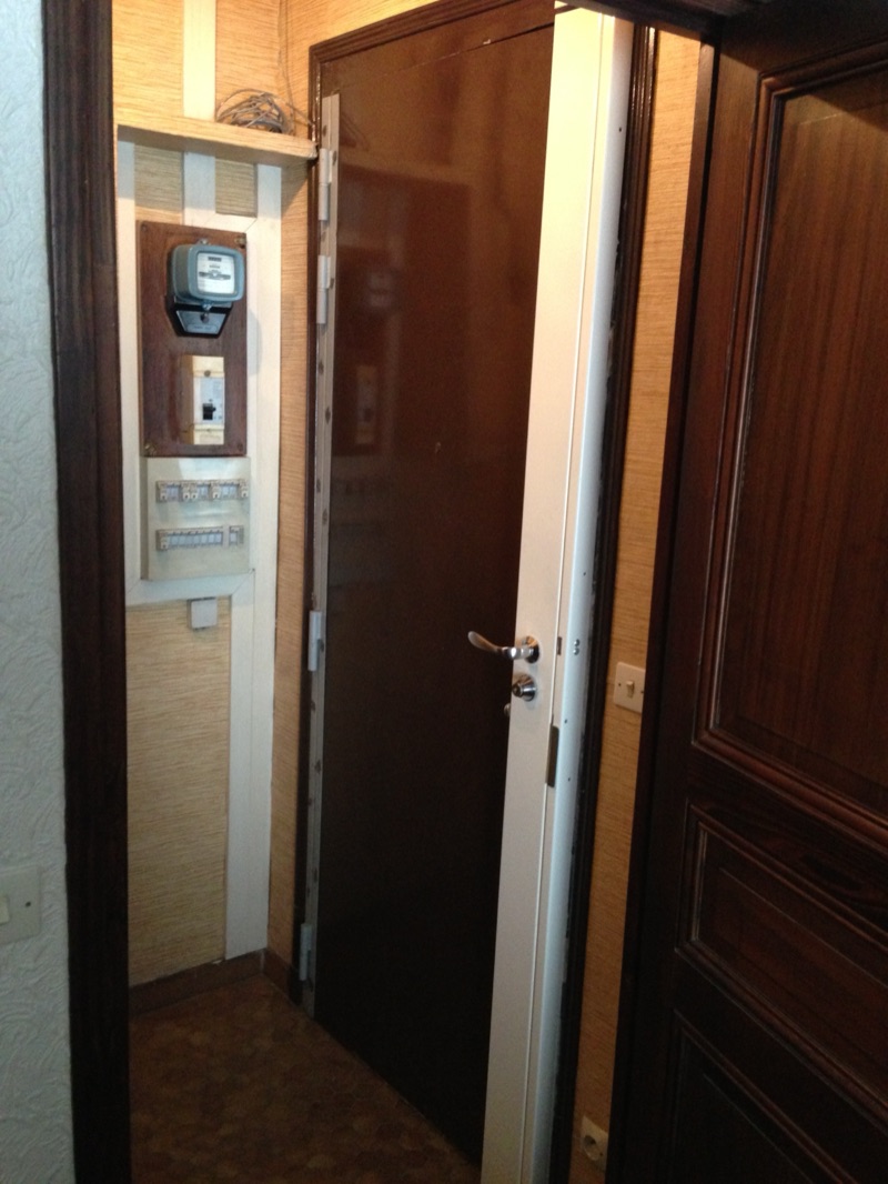 Porte blindée brune entrouverte sur une entrée d'appartement, carénage blanc sur le chant et béquille chromée, tableau électrique à gauche — Paris 15e